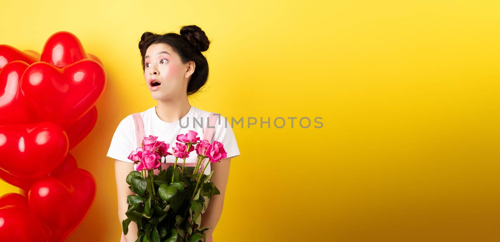 Happy Valentines day. Surprised asian girl looking left with silly face, holding cute pink flowers, receive romantic bouquet, standing on yellow background near red hearts balloons.