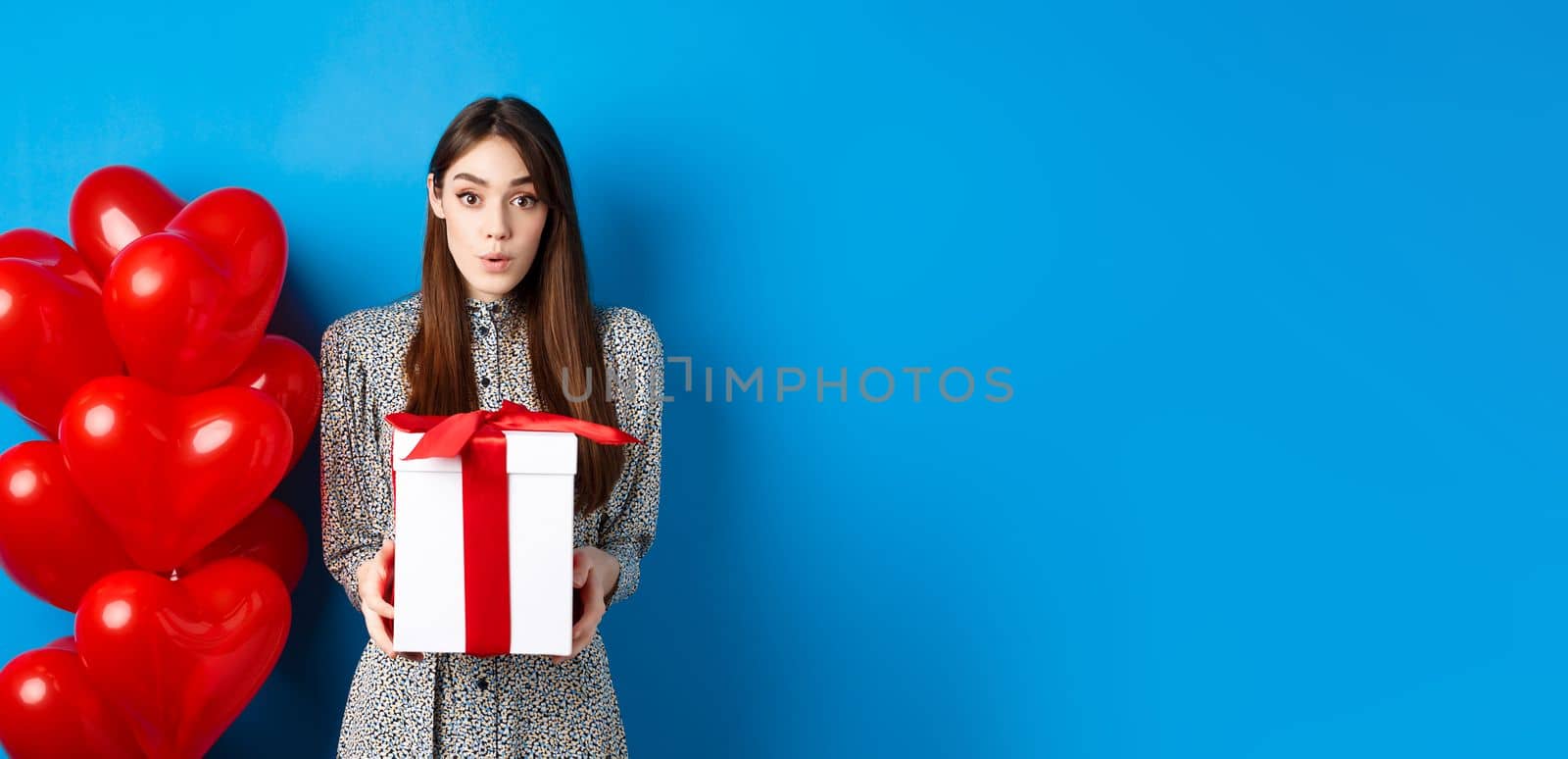 Valentines day. Surprised attractive girl looking amazed at camera, holding big romantic gift, standing near red hearts balloons, blue background.