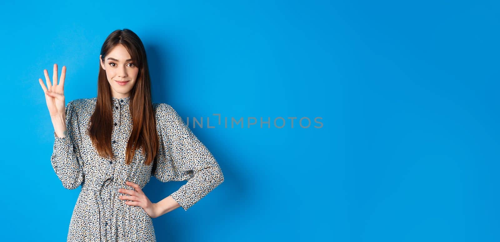 Young beautiful girl in dress with natural long hair, showing number four with fingers and smiling, standing against blue background.