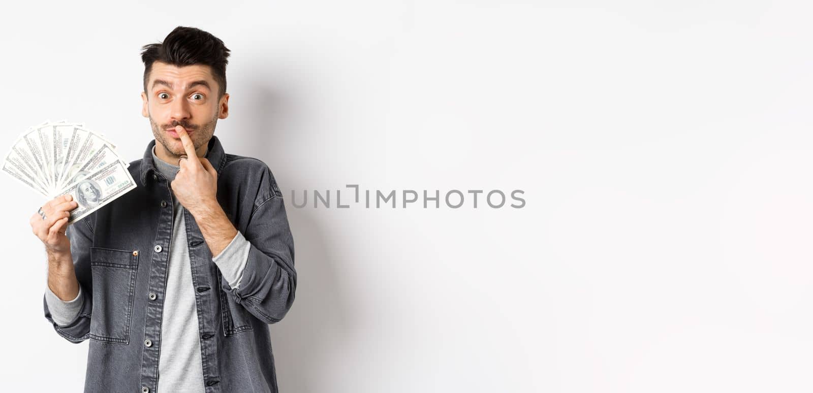 Pensive guy holding money and thinking what buy, pondering about shopping, standing with dollar bills on white background.