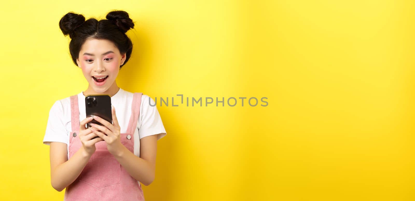 Pretty asian girl looking excited at screen, reading message on phone and smiling happy, standing in summer clothes on yellow background.