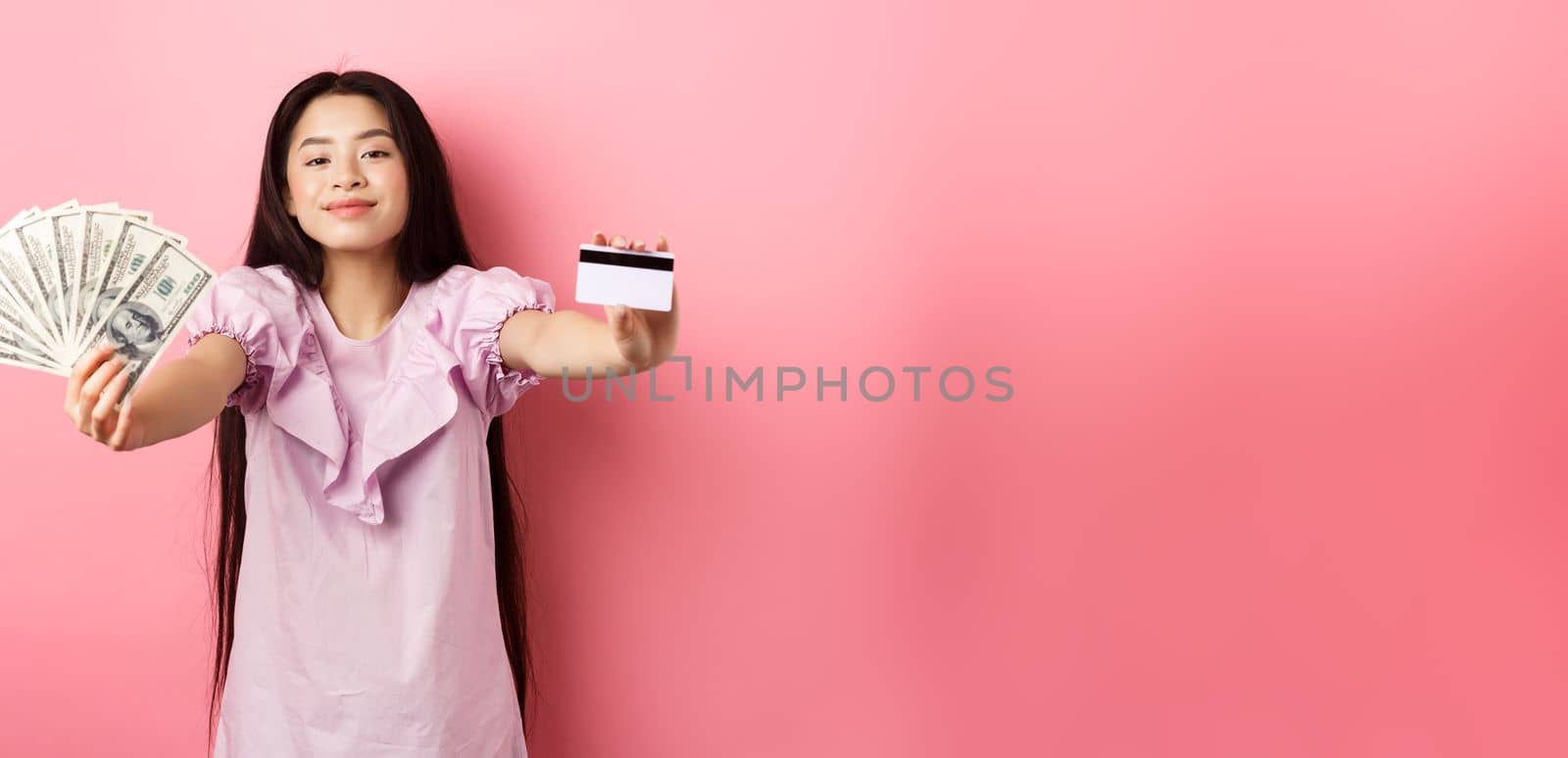 Beautiful asian woman showing plastic credit card and dollar bills, smiling pleased, standing against pink background.