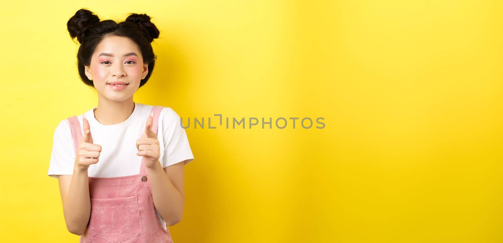 Cheerful teen asian girl pointing fingers at camera and smiling, encourage or invite you, making compliment, praise nice work, standing on yellow background.