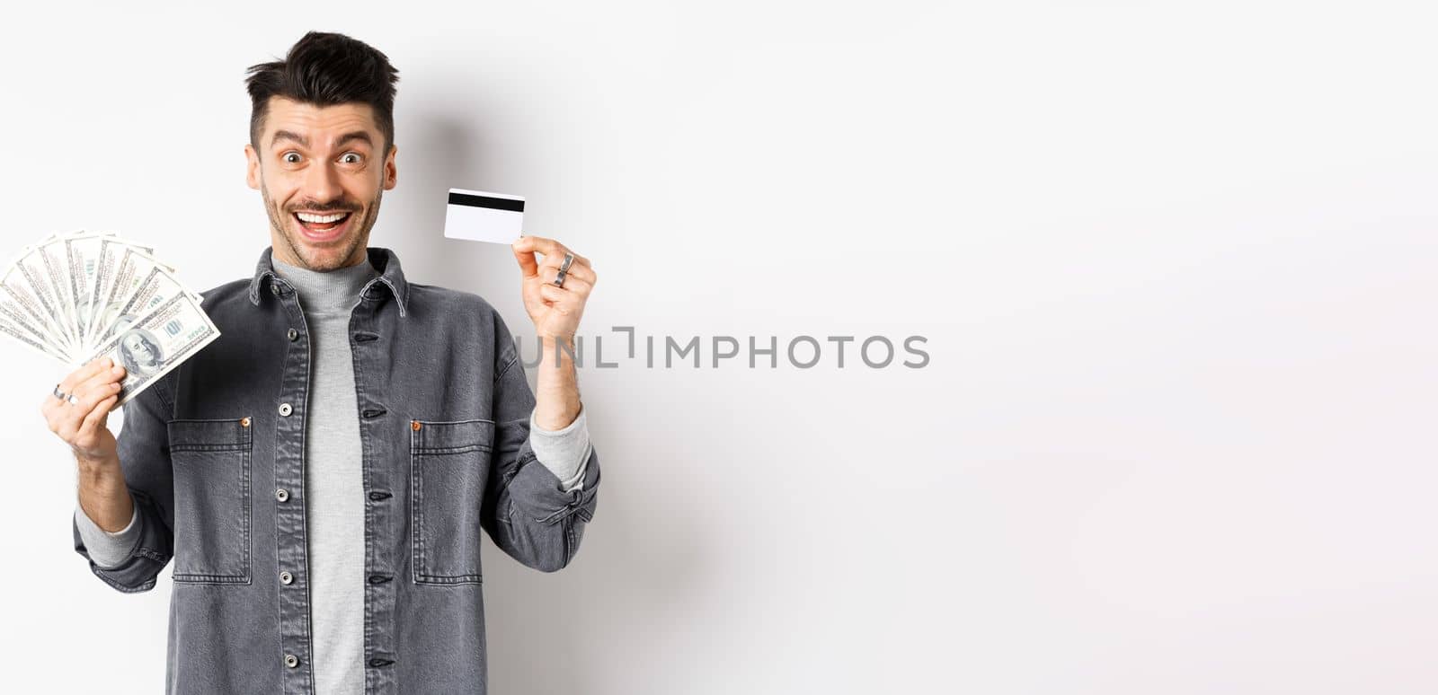 Excited guy holding plastic credit card and dollar bills, standing amused on white background by Benzoix