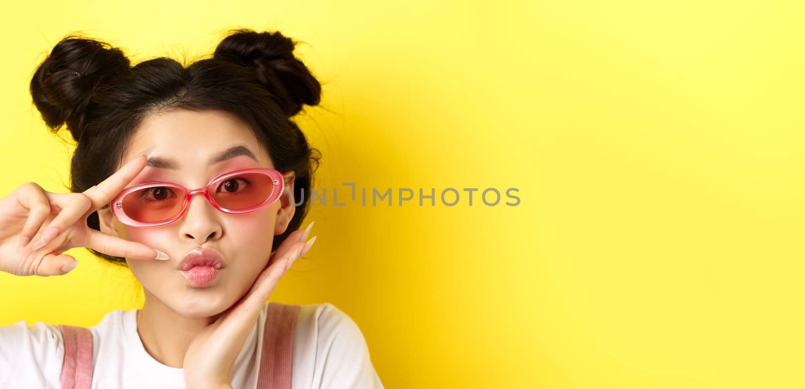 Summer fashion concept. Cute and glamour asian girl in sunglasses, showing v-sign kawaii and looking at camera, standing on yellow background by Benzoix