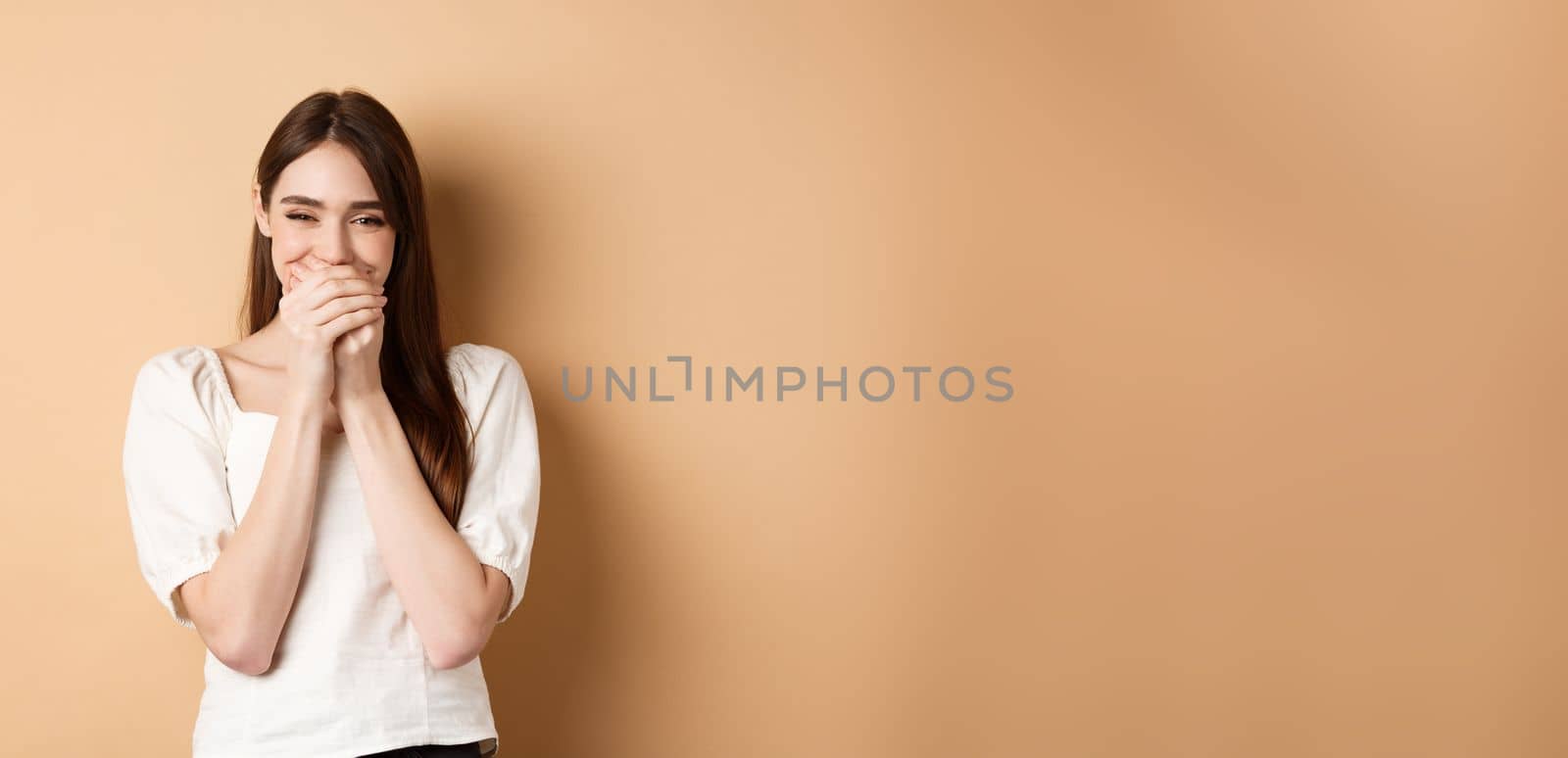 Happy girl laughing and covering mouth with hands, hiding her smile and chuckle over something funny, standing on beige background.