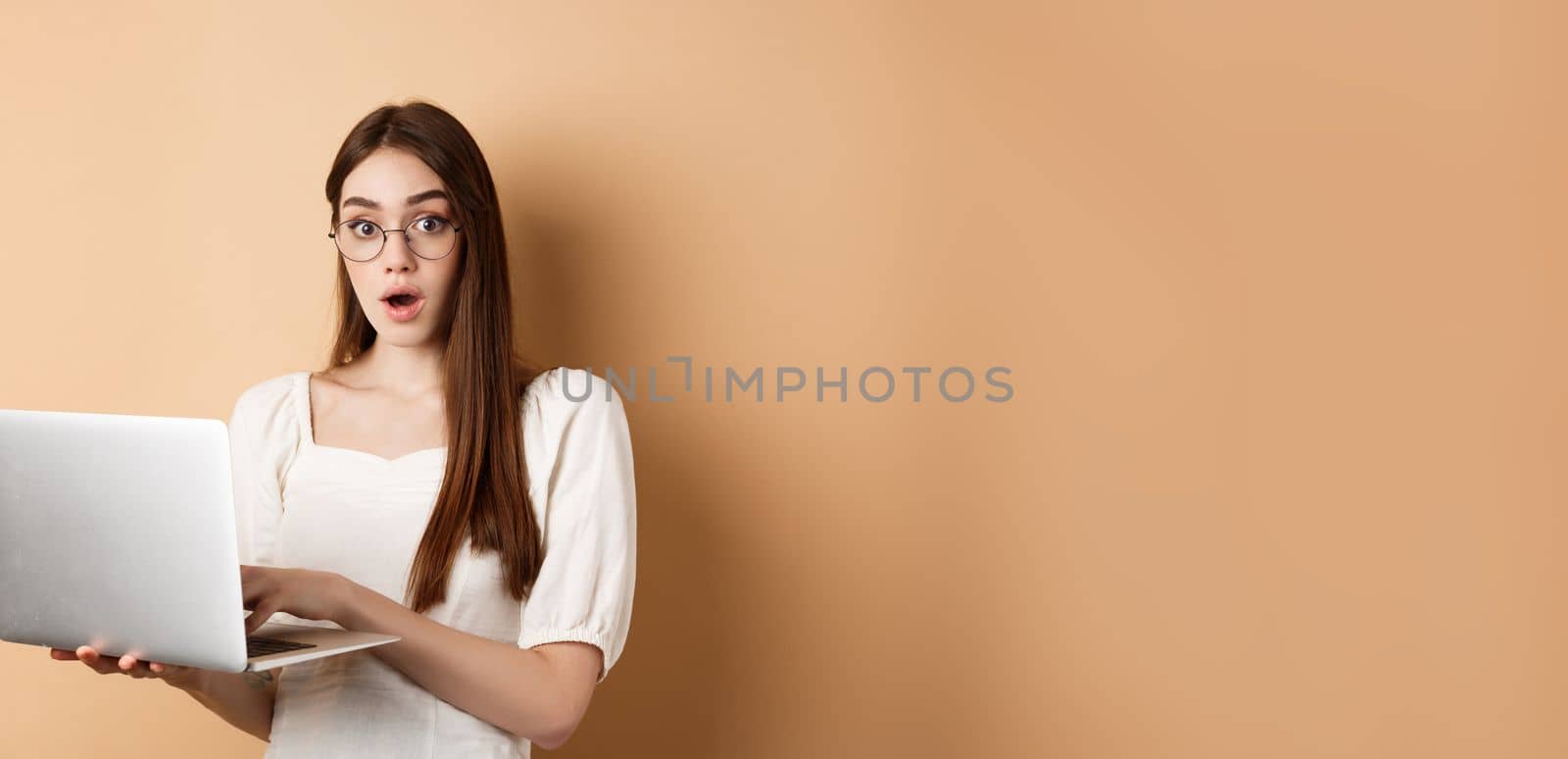 Surprised girl using laptop and gasping amazed, say wow, looking in glasses at camera, beige background.