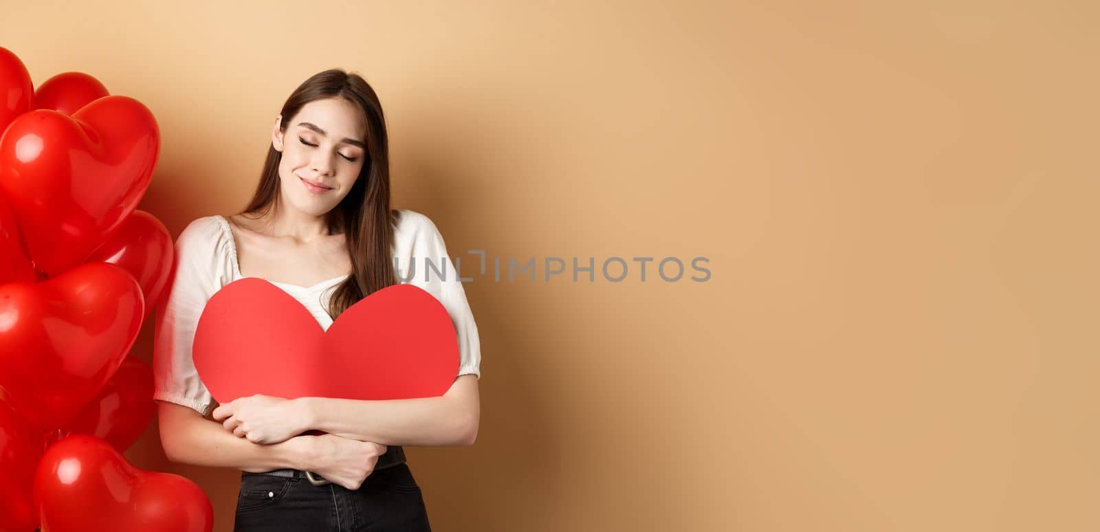 Romantic woman hugging big red heart and smiling dreamy, falling in love on Valentines day, dreaming about lover, standing on beige background near balloons by Benzoix