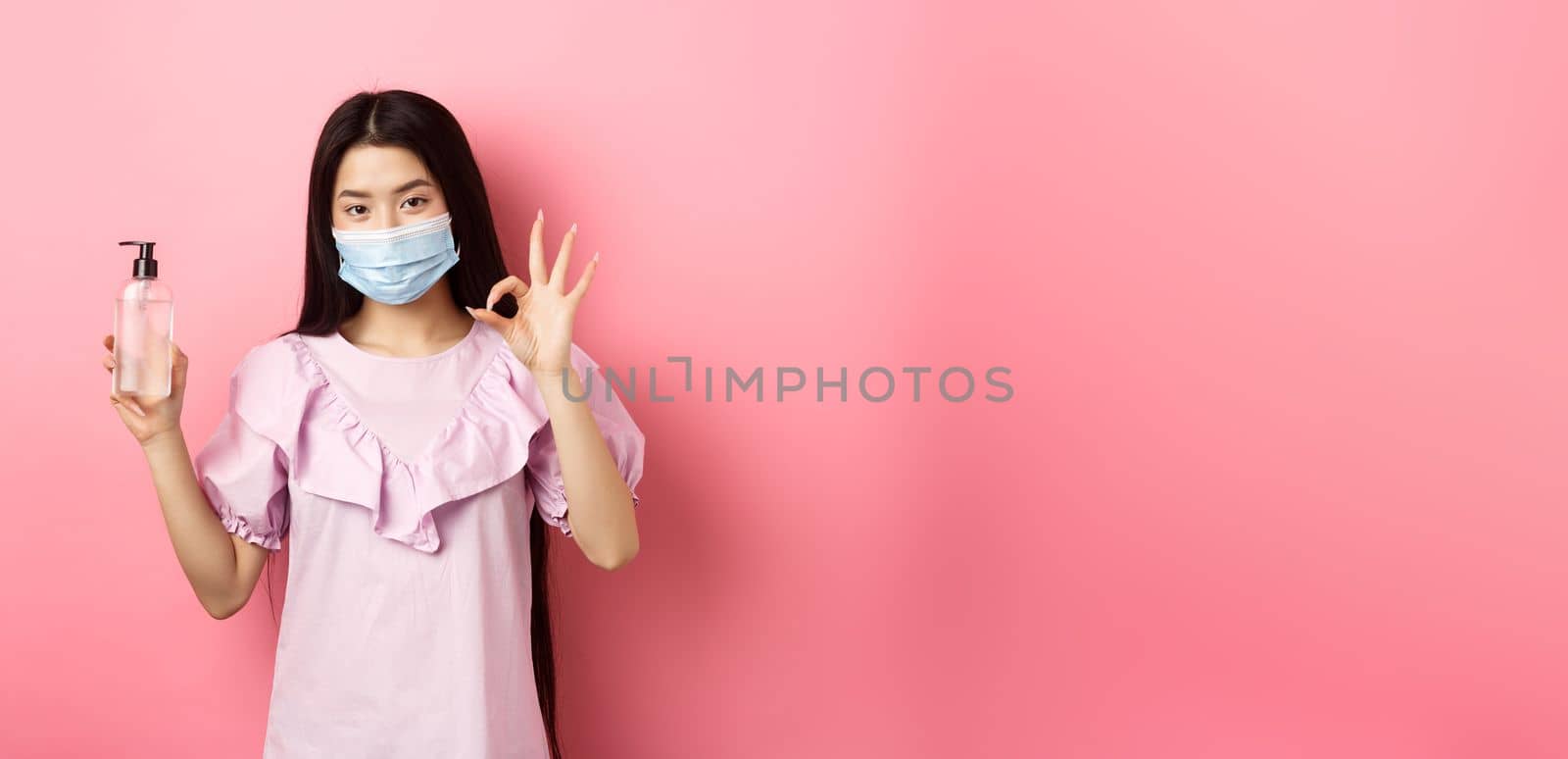 Healthy people and covid-19 pandemic concept. Cheerful asian woman in medical mask recommend hand sanitizer, showing okay sign, good antiseptic bottle, white background by Benzoix