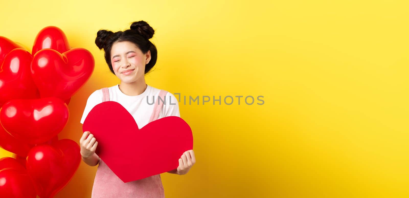 Happy Valentines day. Silly and beautiful asian woman smiling dreamy, showing red heart, imaging romantic date with lover, standing on yellow background by Benzoix