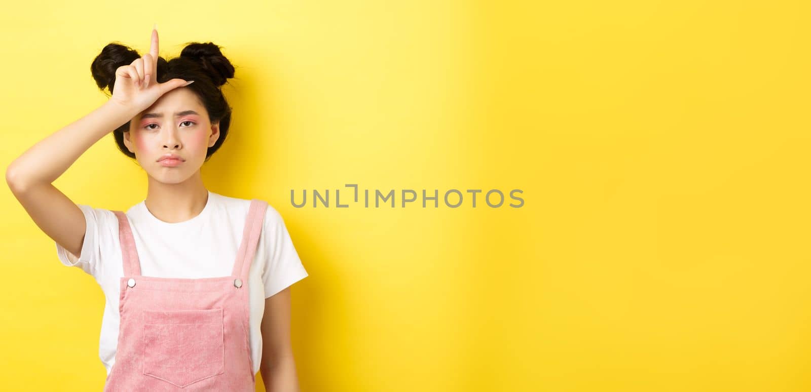 Sad girl showing loser sign on forehead and sulking upset, feeling disappointed in herself, standing on yellow background by Benzoix
