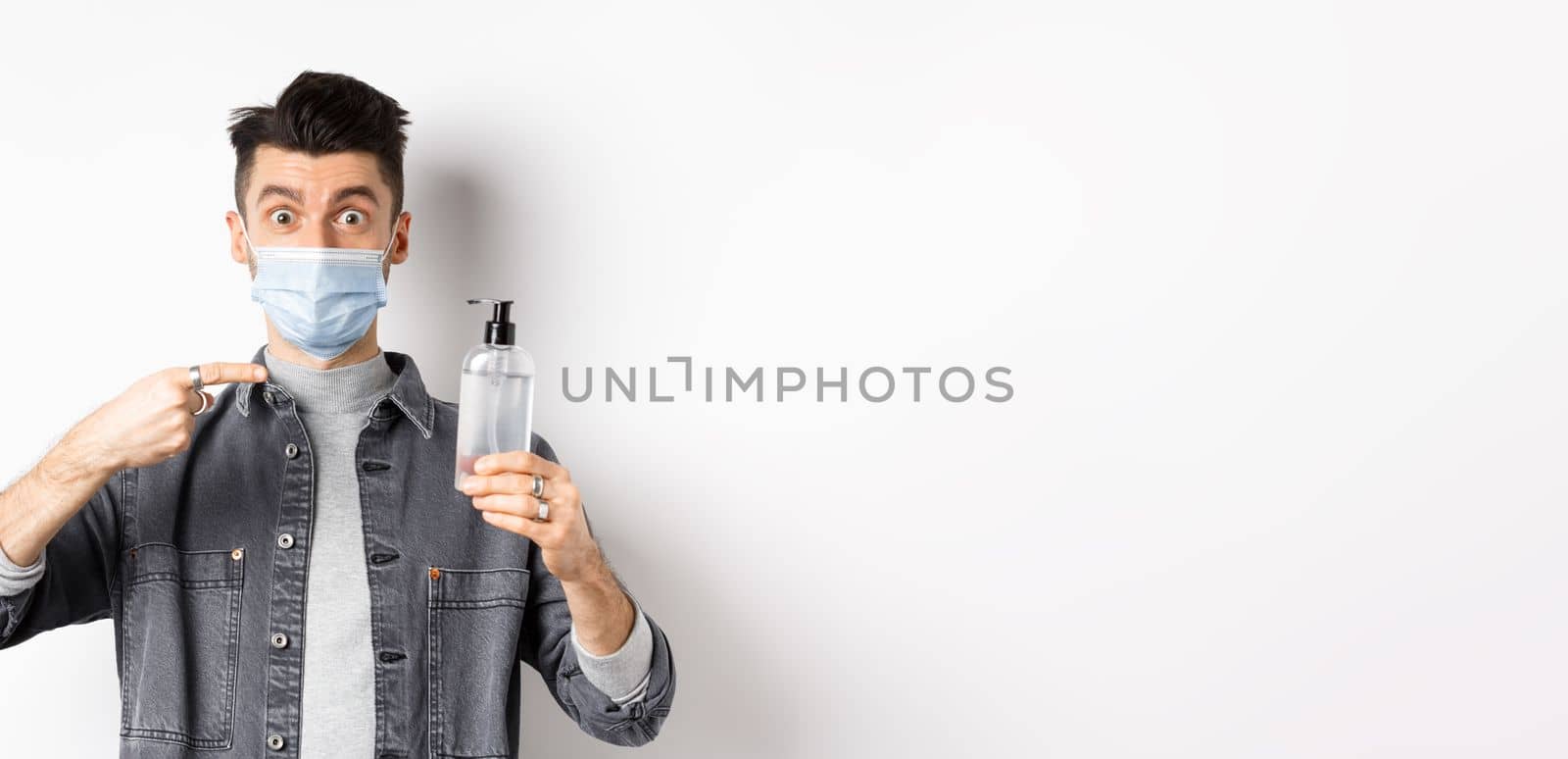 Health, covid and pandemic concept. Stylish guy in medical mask pointing at bottle with hand sanitizer, showing antiseptic, standing on white background.
