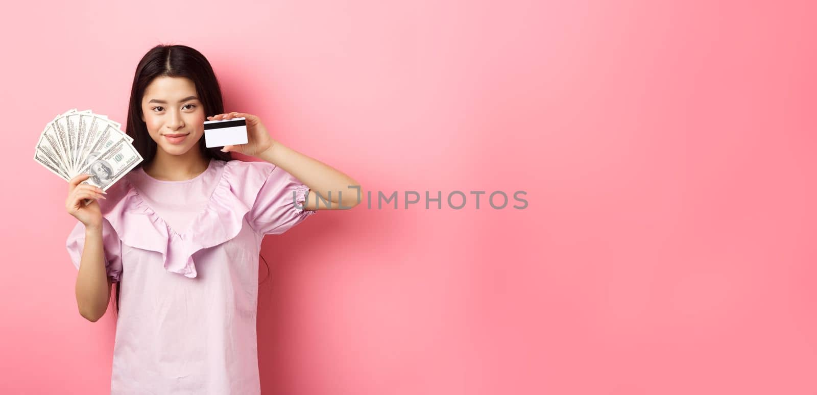 Smiling beautiful asian girl showing money and plastic credit card, giving choice, standing against pink background.