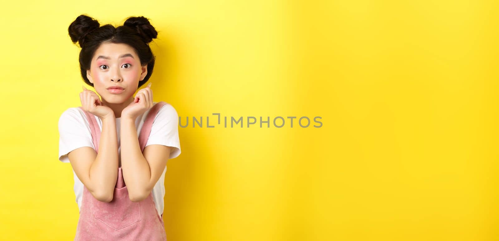 Cute teenage asian girl with beauty makeup, listen with interest, lean on hands and looking excited at camera, standing on yellow background by Benzoix