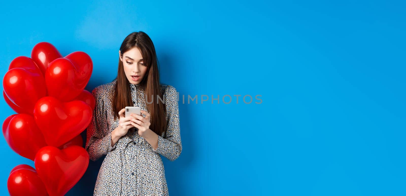 Valentines day. Portrait of young woman standing near red romantic balloons, looking surprised at smartphone screen, blue background by Benzoix