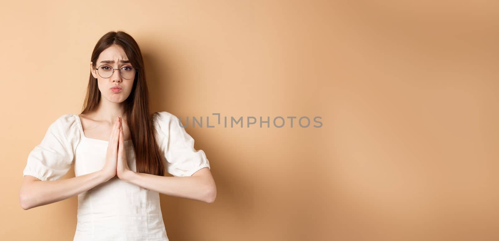 Sad girl in glasses begging for help, say please and looking cute, need favour, standing on beige background.