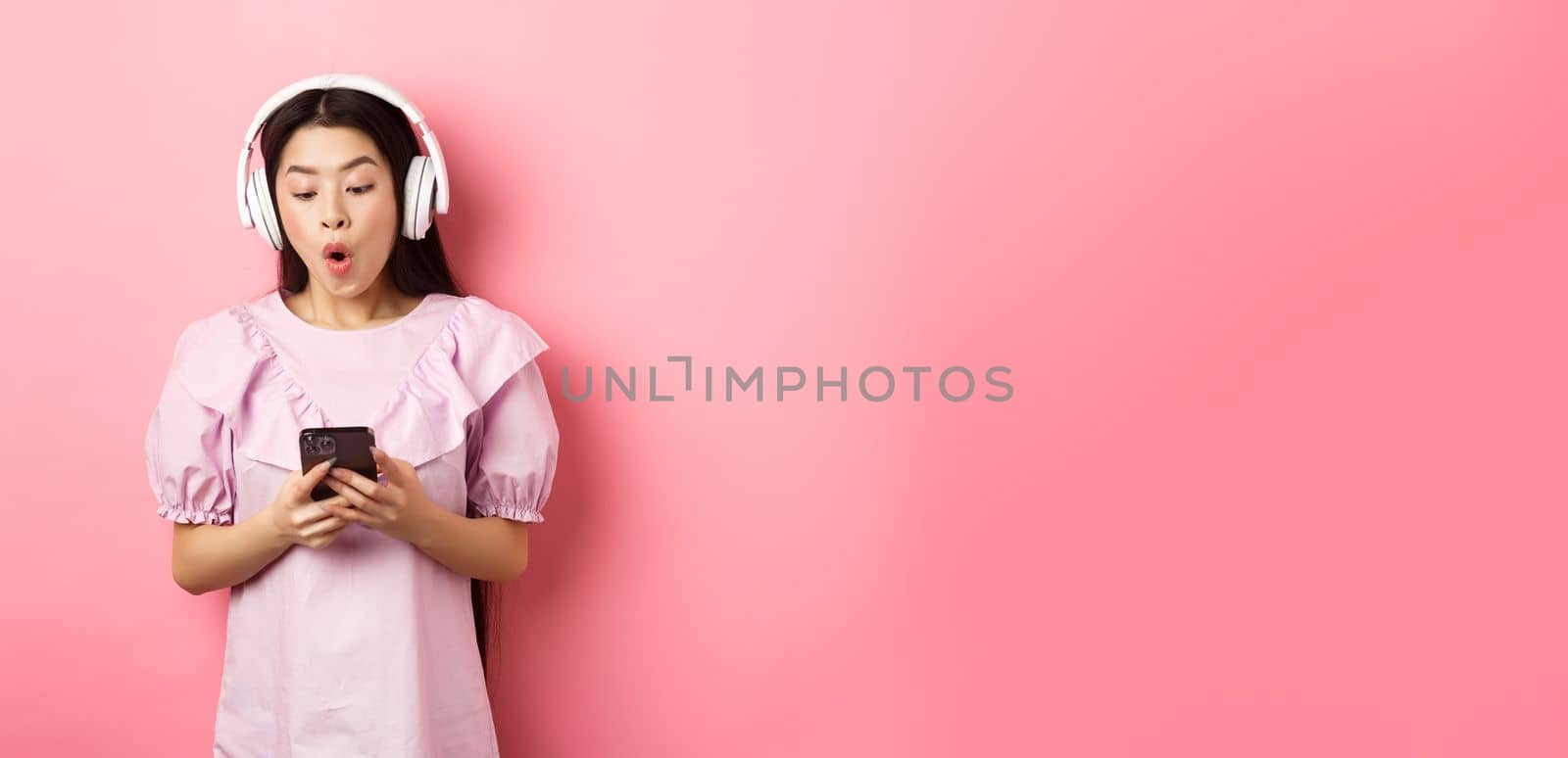 Excited asian girl looking at smartphone screen amused, say wow, listening music in wireless headphones, standing against pink background.