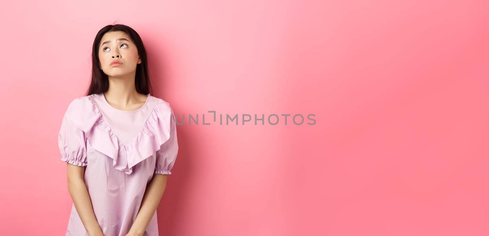 Dreamy and sad asian woman look at upper left corner logo, standing pensive and gloomy in pink romantic dress, white background by Benzoix