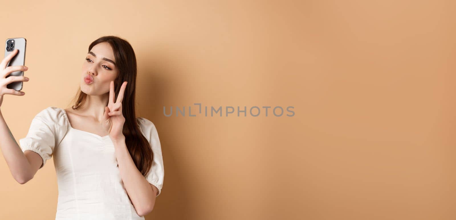 Fashion female blogger pucker lips and show v-sign at smartphone camera, taking selfie for social media, standing on beige background.