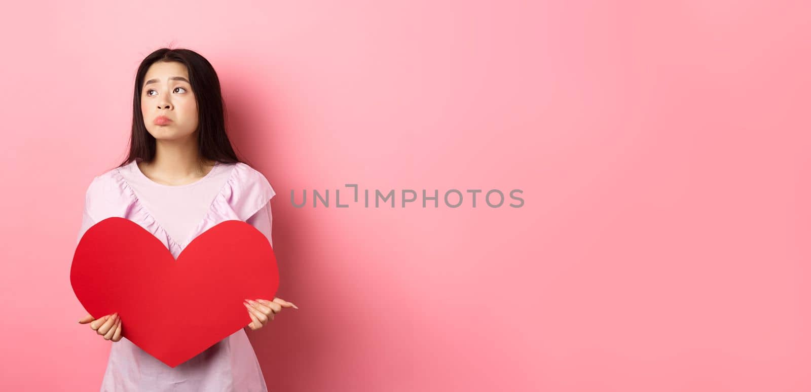 Valentines concept. Lonely teenage asian girl dreaming about love, feeling sad and lonely on lovers day, looking aside with pity, holding big red heart, pink background.
