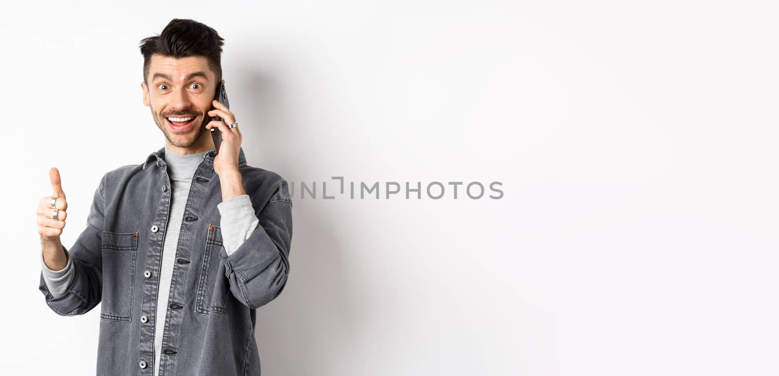 Excited young man talking on phone and showing thumbs up, smiling satisfied, standing against white background.