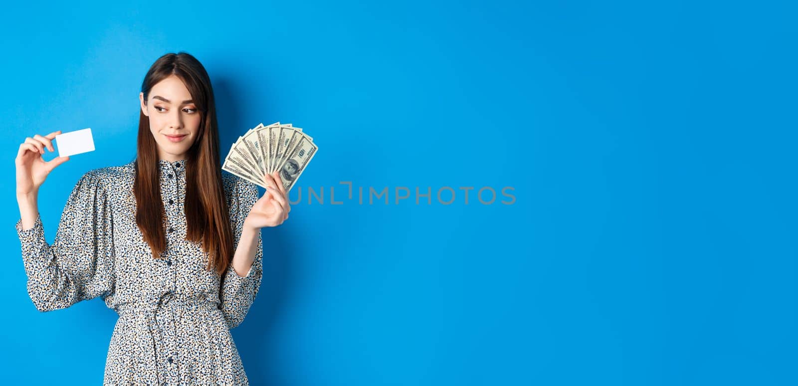 Shopping. Beauty girl in dress looking aside and smiling pensive, showing money and plastic credit card, standing on blue background by Benzoix