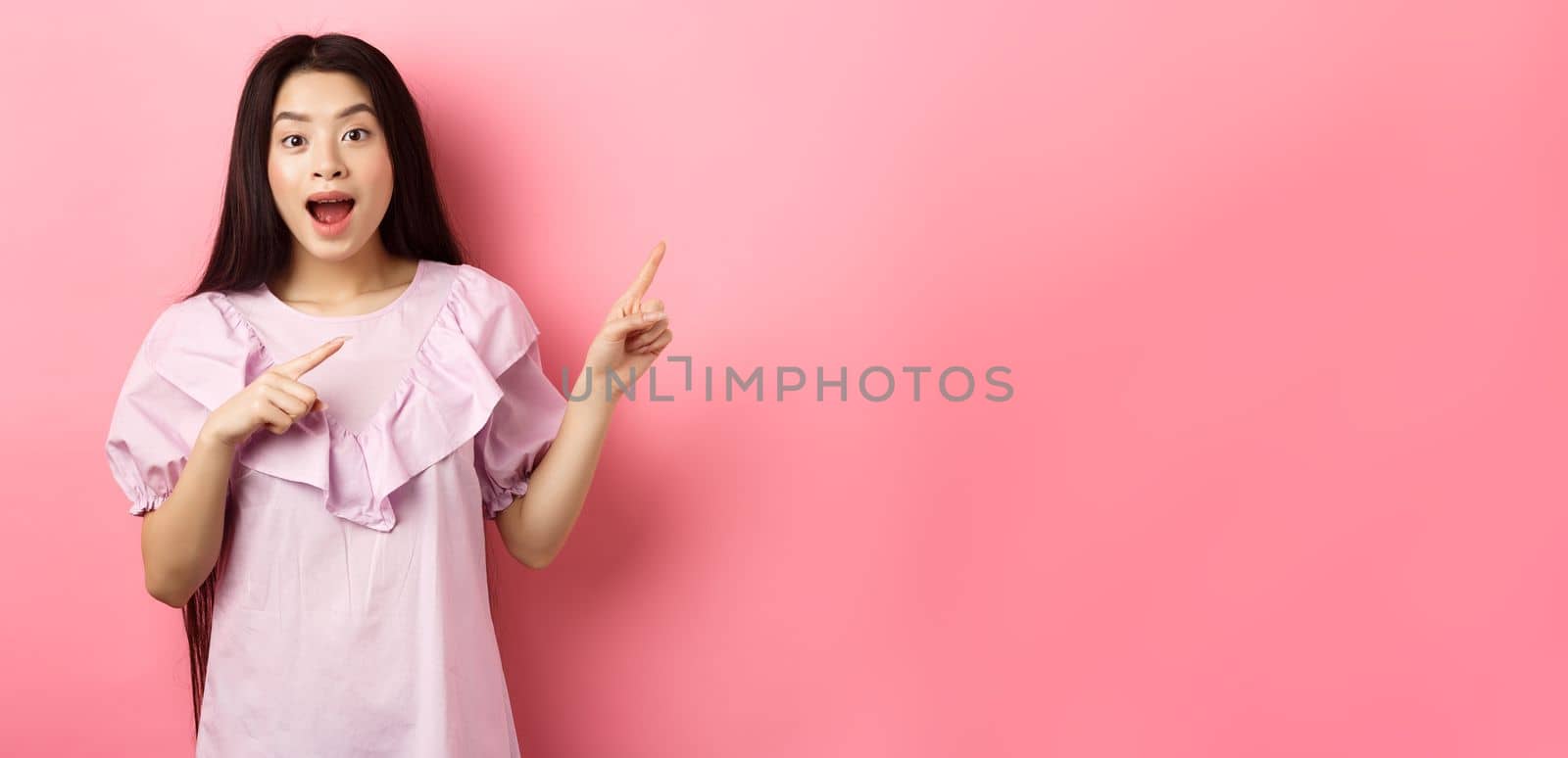 Excited asian teenage girl pointing fingers right, smiling happy and amused, showing cool news, standing on pink background by Benzoix
