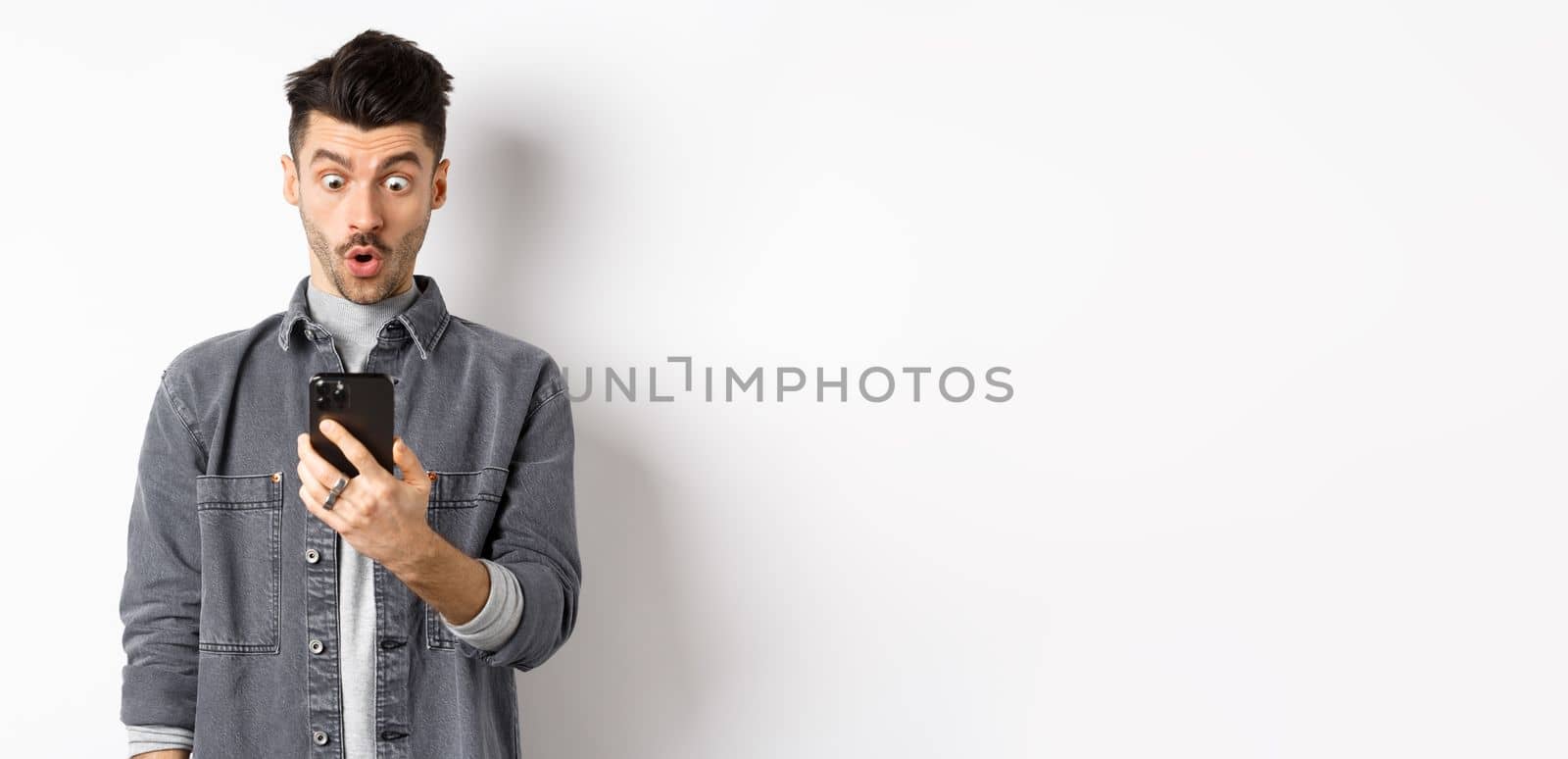 Man look surprised at smartphone screen, receive exciting news on phone, standing against white background by Benzoix