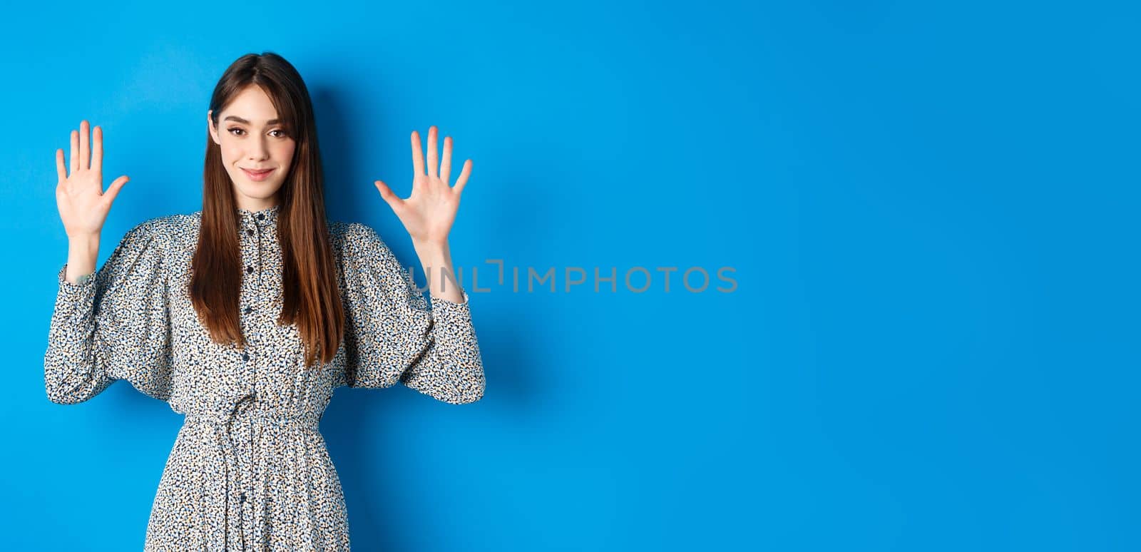 Young beautiful girl in dress with natural long hair, showing number ten with fingers and smiling, standing against blue background.