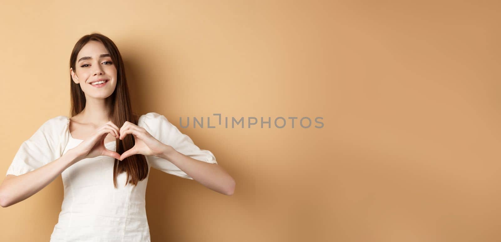 Valentines day. Attractive young girl in white dress smiling and showing heart gesture, standing on beige background. Copy space