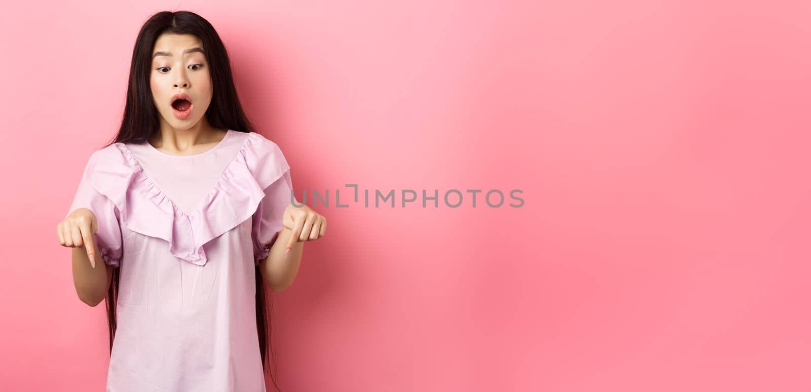 Surprised asian woman in awe, drop jaw and gasping, pointing fingers down at amazing promo offer, standing impressed against pink background by Benzoix