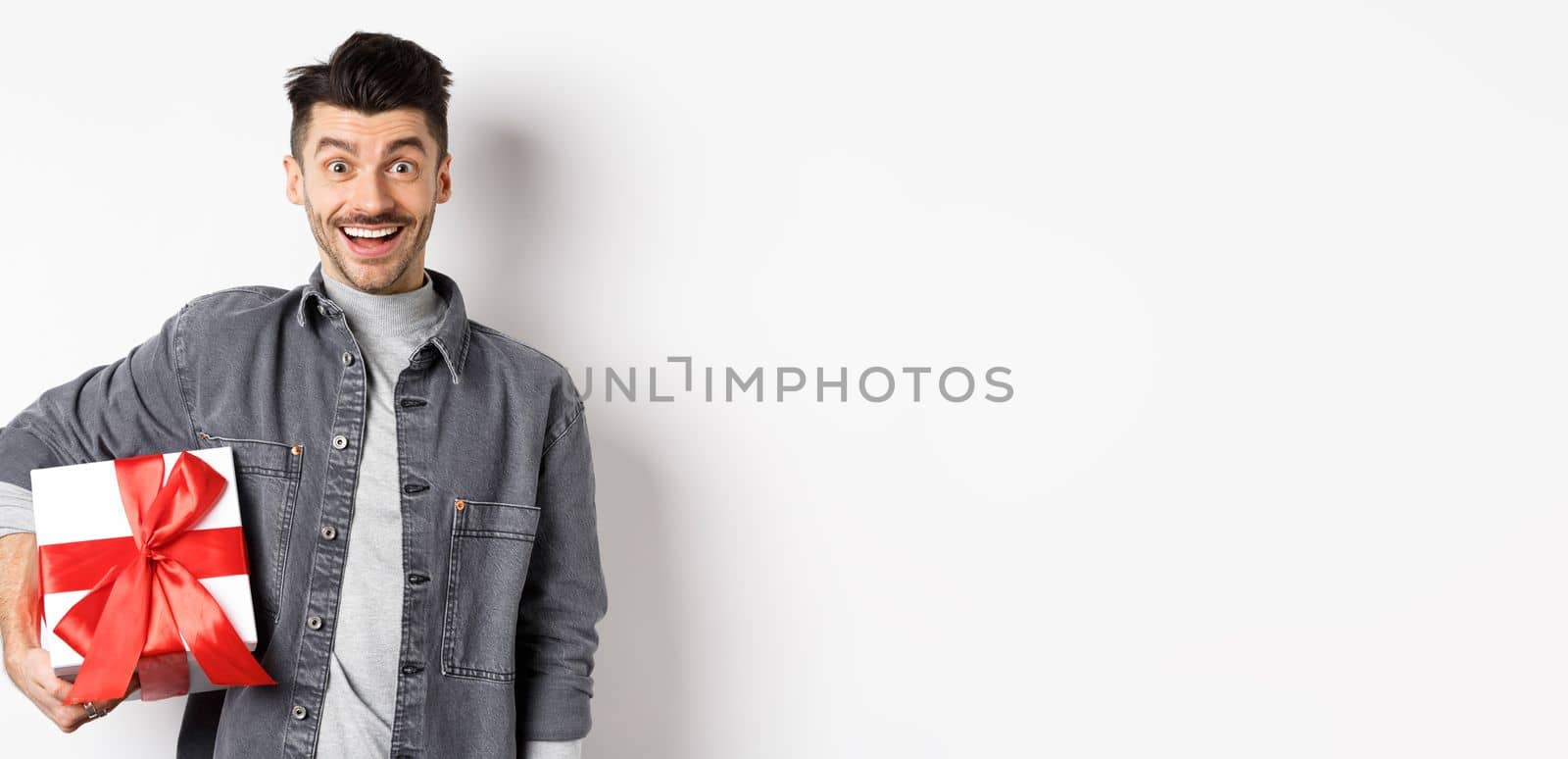 Happy Valentines day. Surprised and happy guy holding gift box and look at camera, smiling amazed, celebrating holiday, bring present on romantic date, white background.