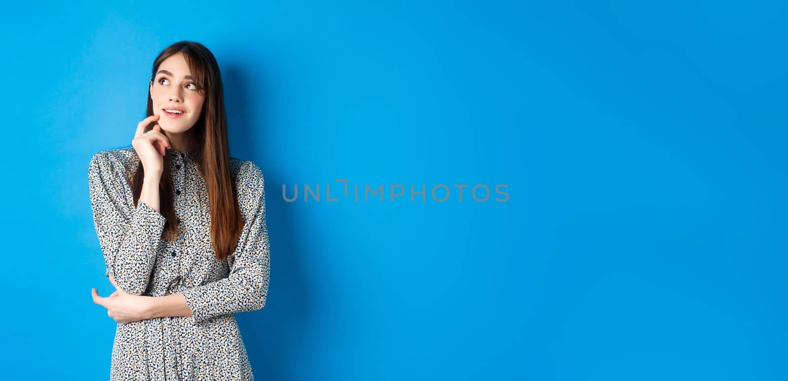 Creative young woman imaging and smiling, looking at upper left logo thoughtful, having an idea, standing on blue background in dress by Benzoix
