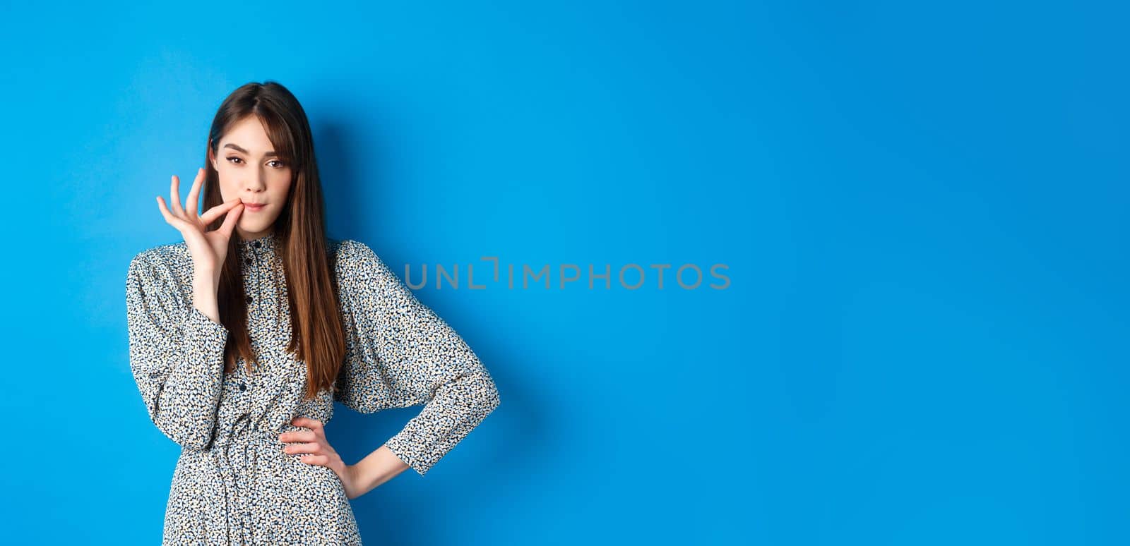 Serious woman promise to keep secret, seal lips, making zip gesture and looking at camera, standing in dress on blue background.