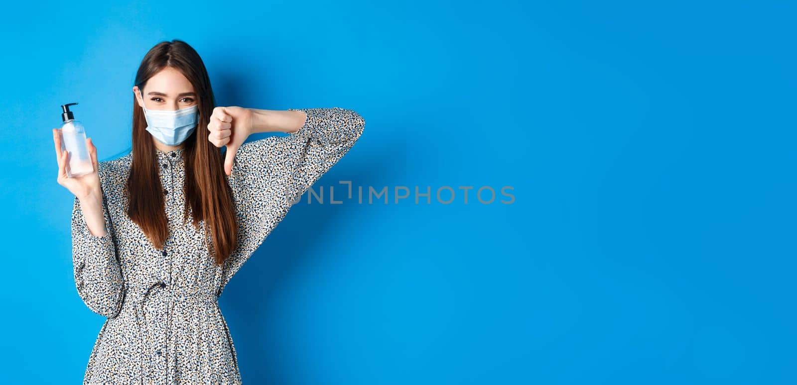 Covid-19, social distancing and healthcare concept. Disappointed girl in medical mask showing bad hand sanitizer and thumbs down, standing on blue background.