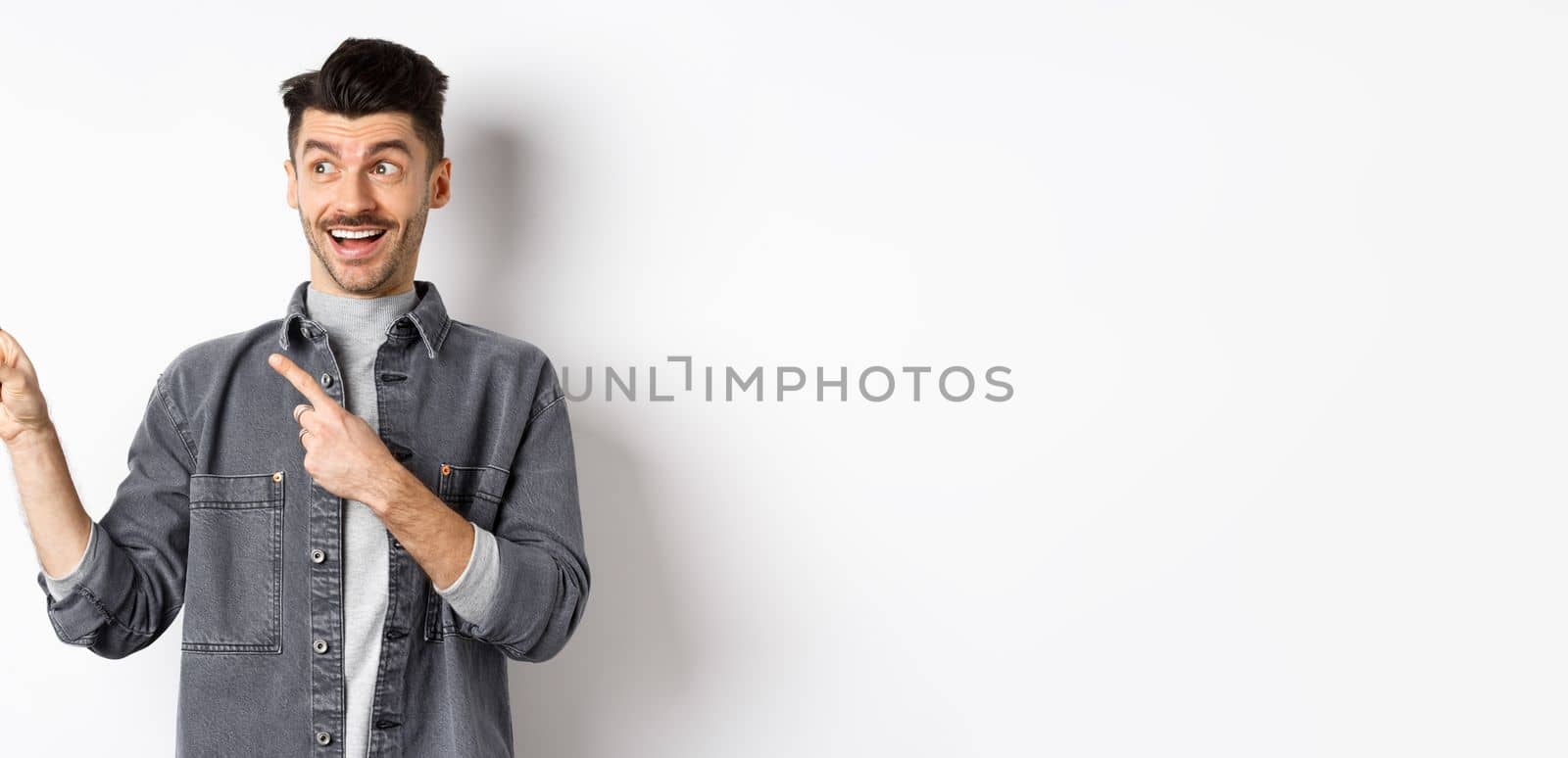 Handsome smiling guy checking out advertisement, looking and pointing left with excited face, standing on white background.