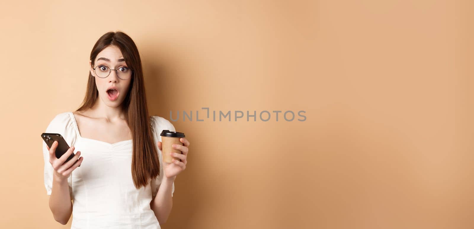 Surprised girl react to mobile phone news, holding smartphone and cup of coffee, look excited with dropped jaw at camera, beige background.