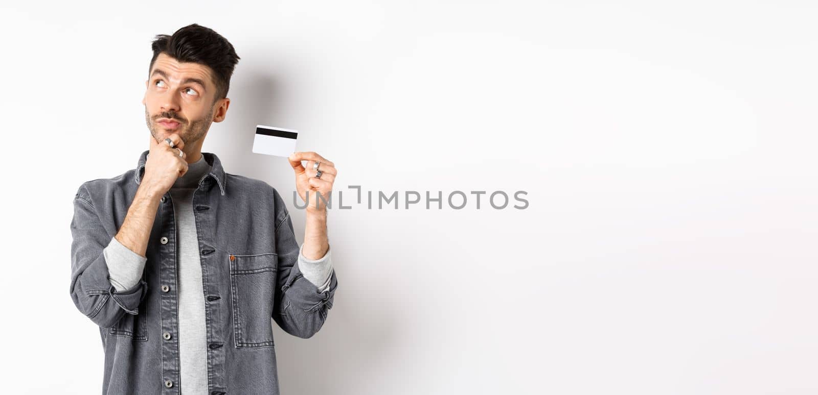 Thoughtful guy looking at upper left corner logo and holding plastic credit card, thinking about shopping, standing on white background.
