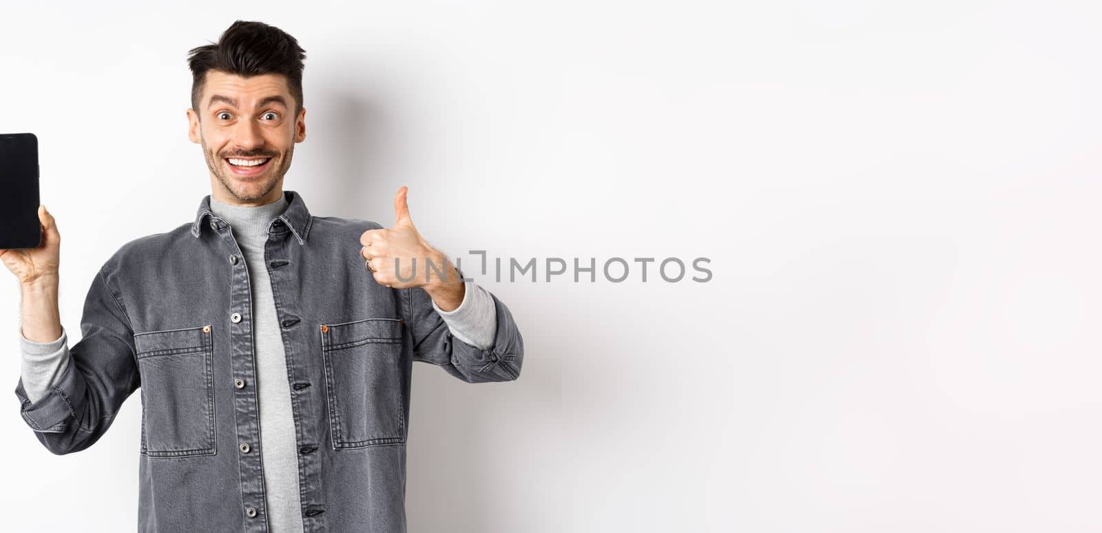 Cheerful stylish man showing empty smartphone screen and thumbs-up, concept of online shopping and e-commerce.