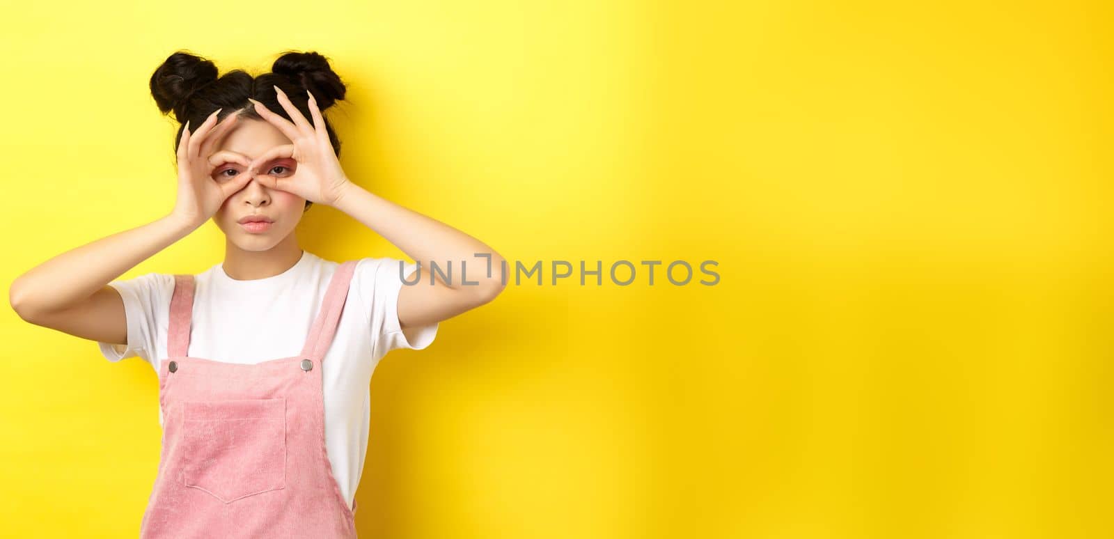 Funny asian girl look through hand binoculars, watching you, standing in summer clothes on yellow background.