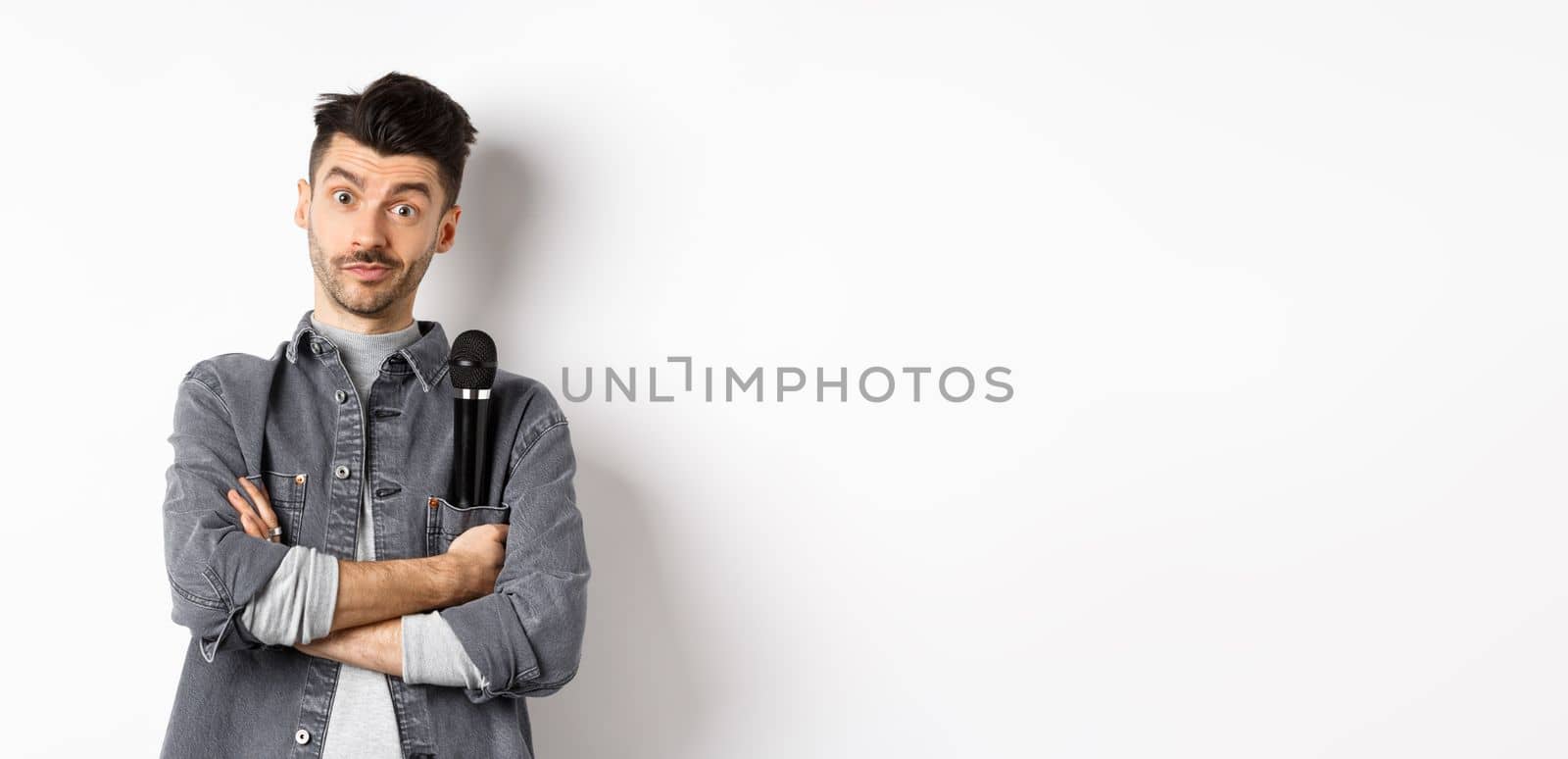 Image of funny male entertainer or singer, holding microphone in pocket of jacket and look at camera with arms crossed, standing against white background.
