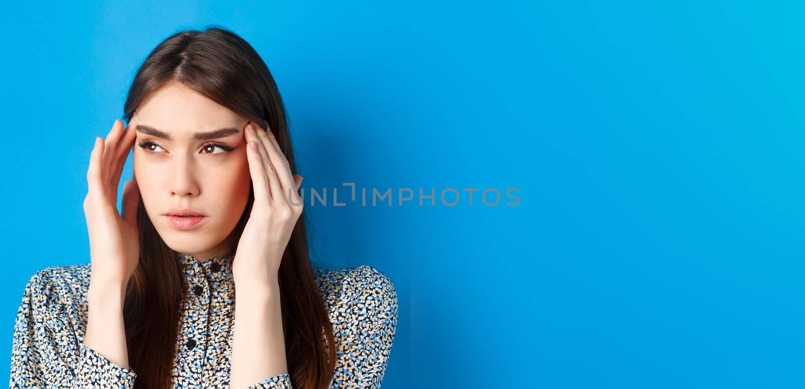 Close-up portrait of young woman feel sick, touching head temples and frowning from headache, having migraine, looking aside at logo, blue background.