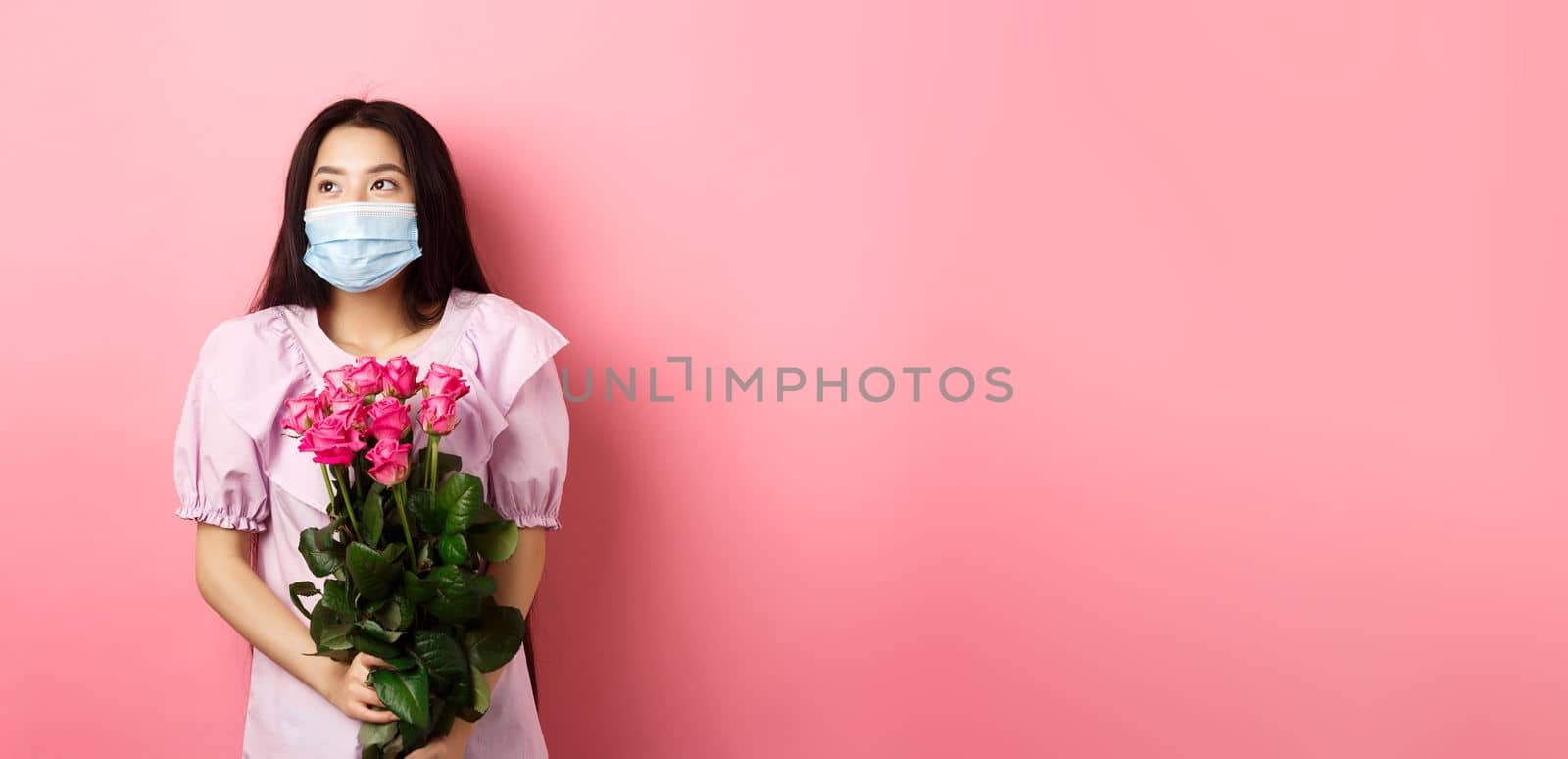 Romantic asian girl in medical mask looking aside at empty space with dreamy eyes, holding bouquet of roses for Valentines day, having date with lover during pandemic.