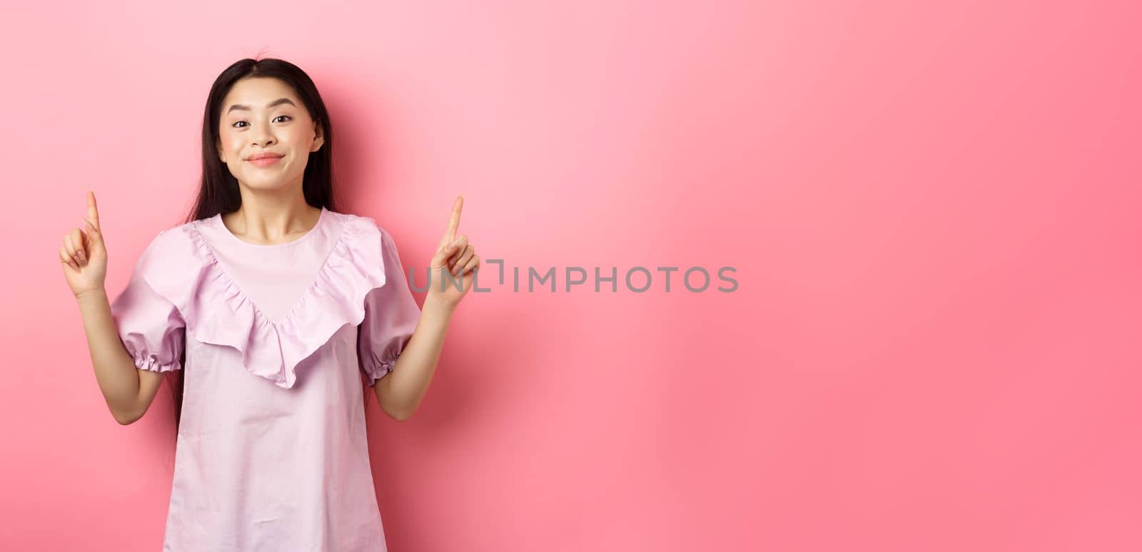 Beautiful young asian woman in romantic dress pointing fingers up, smiling and gladly showing advertisement, standing on pink background by Benzoix