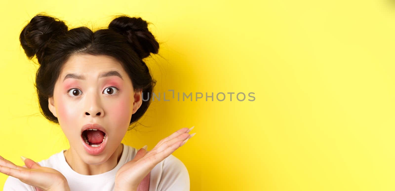 Close-up of excited asian woman with glamour makeup and hairstyle, screaming of amazement, open mouth and look at camera in awe, yellow background.