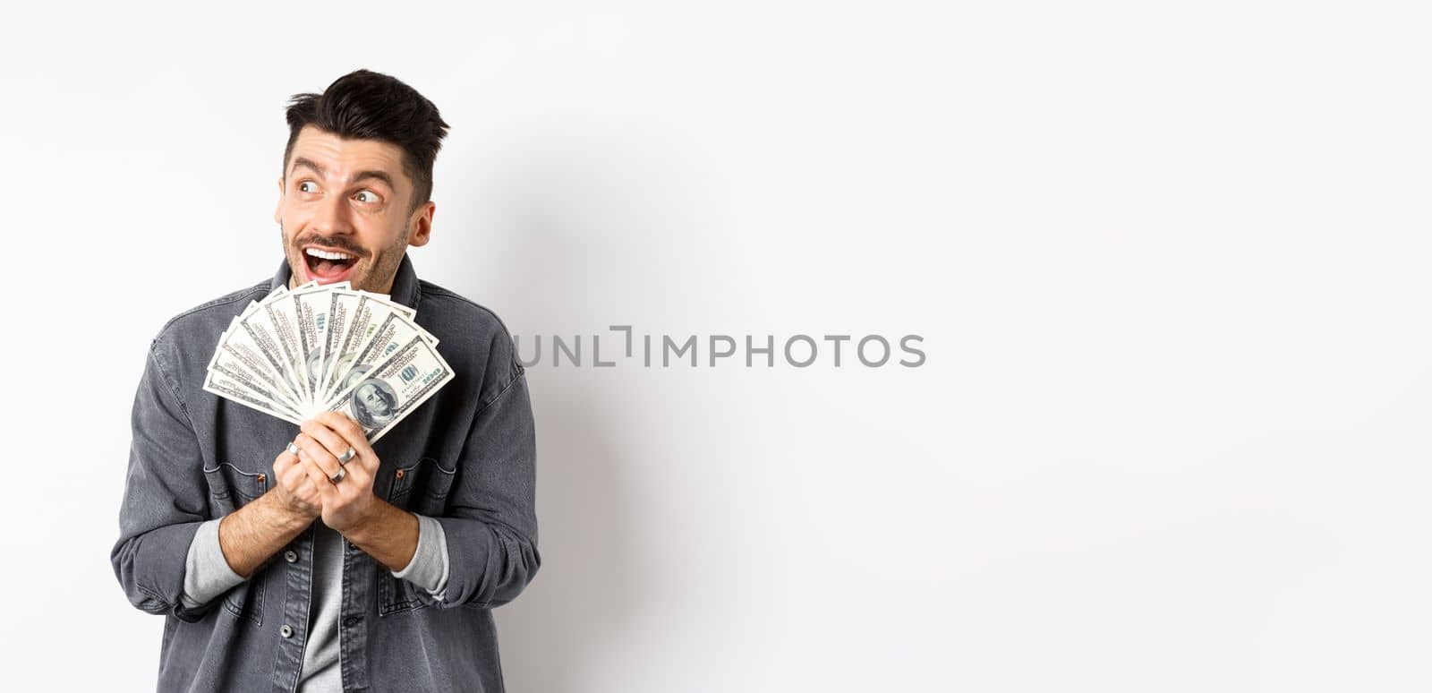 Rich happy man dreaming of shopping wish dollar bills in hands, looking aside pensive and holding money, standing against white background.