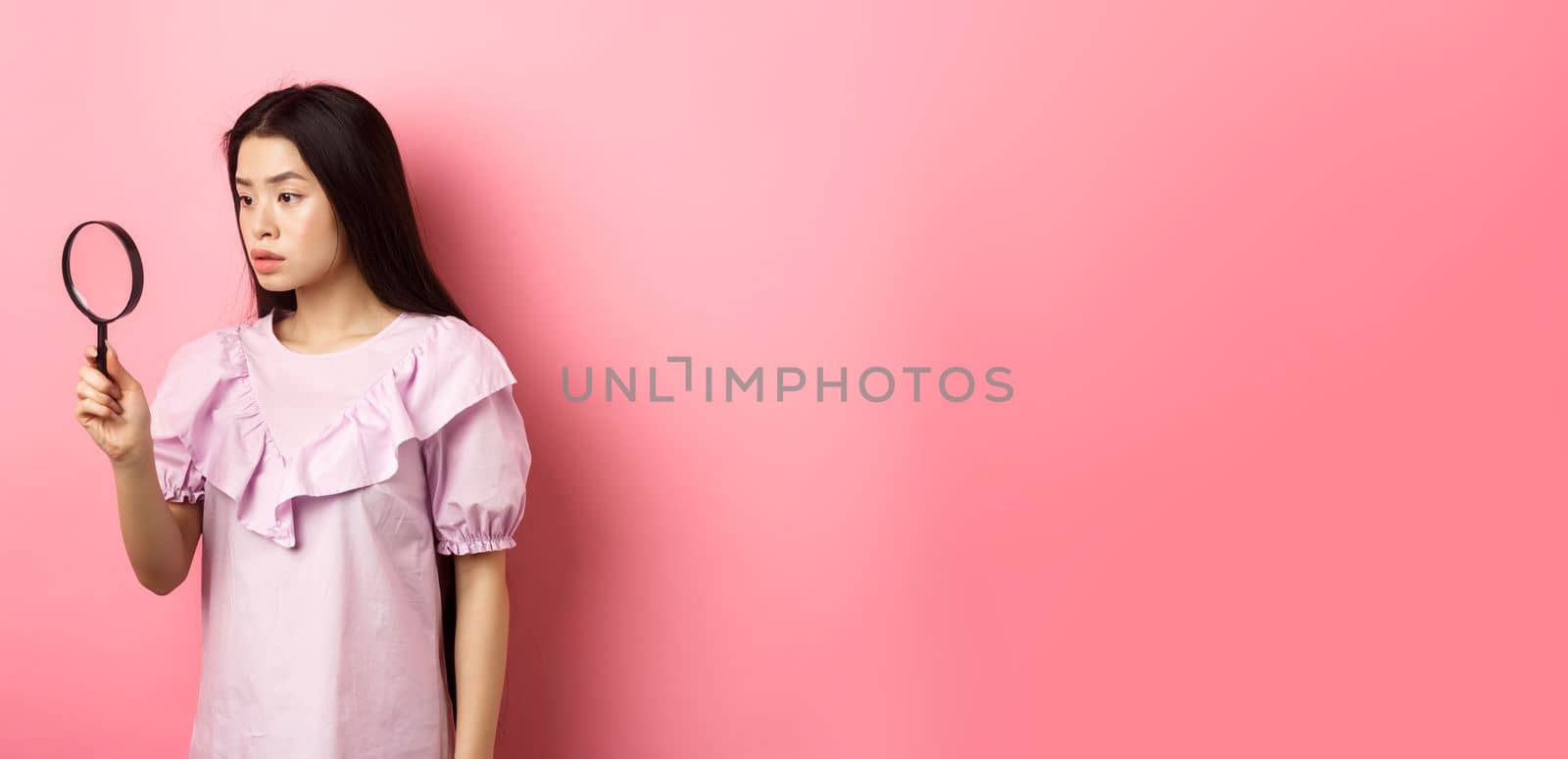 Serious girl investigating, looking through magnifying glass, searching for something, standing against pink background.