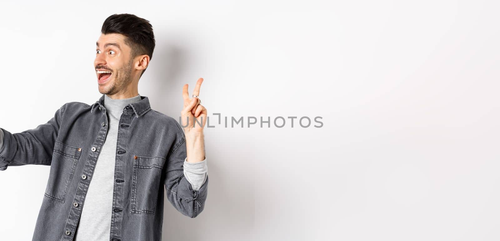 Profile of cheerful young guy taking selfie on smartphone, showing v-sign while make photo on mobile app, standing against white background by Benzoix