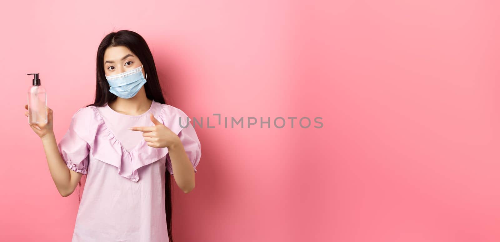 Healthy people and covid-19 pandemic concept. Stylish japanese girl in medical mask showing bottle of hand sanitizer, pointing at antiseptic, standing against pink background by Benzoix