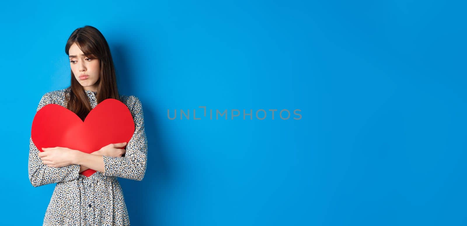 Valentines day. Lonely single girl hugging big red heart cutout and looking down upset, standing gloomy on blue background by Benzoix